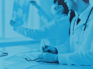 Picture of a doctor holding an x-ray. Doctor is also wearing a medical mask and white lab coat. The doctor to the left is wearing a white lab coat, stethoscope and writing on clipboard. Image has a blue overlay.