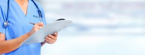 Picture of nurse wearing blue scrubs, a stethoscope around neck and holding a clipboard while writing