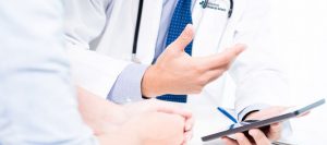 Picture of doctor talking to patient. Doctor is holding a phone and showing it to the patient. Doctor is wearing a white lab coat and a blue dotted tie. Patient is wearing a very light blue shirt.