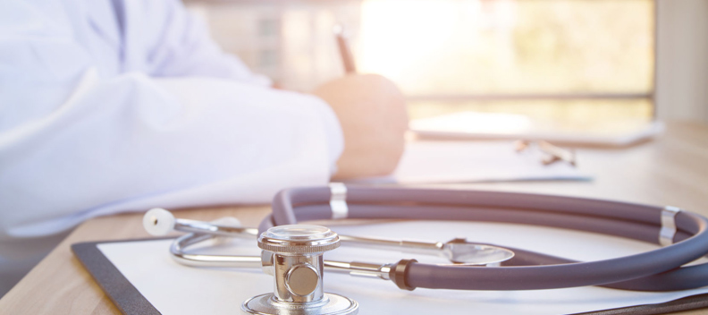 Doctor sitting at a desk writing on a clipboard with a stethoscope and some paperwork on the desk.