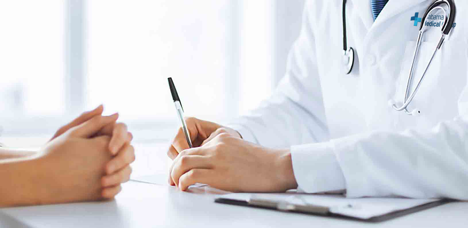 Doctor sitting across a table from a patient. Doctor is holding a pen and arm is cover a small clipboard of paper.