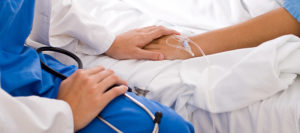Person in blue scrubs holding a stethoscope and a patients hand. Patient is laying in bed with an iv in the hand the person is holding.